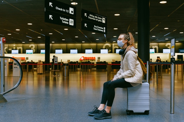 ragazza bionda con occhiali e mascherina seduta su valigia in aeroporto vuoto