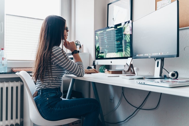 raggaza con capelli lunghi seduta a scrivania lavora con due monitor del computer